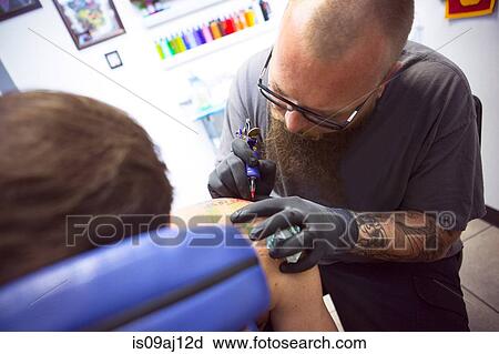 Stock Image - Mid adult man having shoulder tattooed in tattoo parlor. Fotosearch