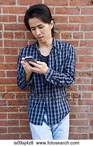 Stock Photo - Mid adult man leaning against wall texting on smartphone. Fotosearch