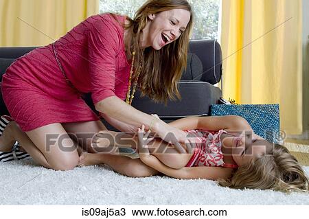 Stock Image - Mid adult mother tickling daughter on sitting room rug. Fotosearch