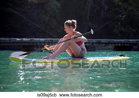 Stock Image - Mid adult woman sit down paddleboarding at sea. Fotosearch