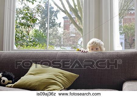 Stock Photo - Portrait of cute male blond toddler hiding behind sofa. Fotosearch