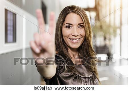 Stock Image - Portrait of mid adult businesswoman holding up hand in victory sign. Fotosearch
