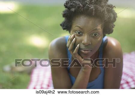 Stock Image - Portrait of young woman lying on picnic blanket in park. Fotosearch