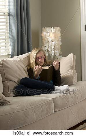 Stock Image - Pre adolescent girl reclining on sofa reading book. Fotosearch