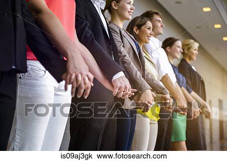 Stock Image - Row of businesswomen and men holding hands in office. Fotosearch