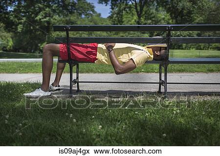 Stock Photo - Side view of young man lying on park bench with fedora covering face. Fotosearch
