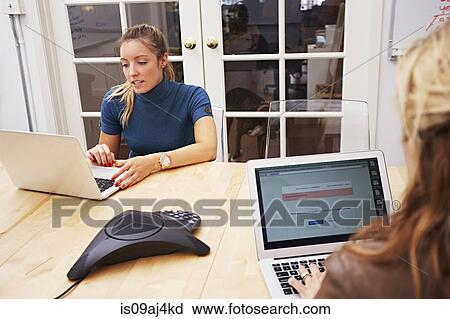 Stock Image - Two female colleagues using laptops in office. Fotosearch