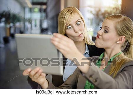 Stock Image - Two young businesswomen pulling faces and taking selfie on digital tablet in office. Fotosearch