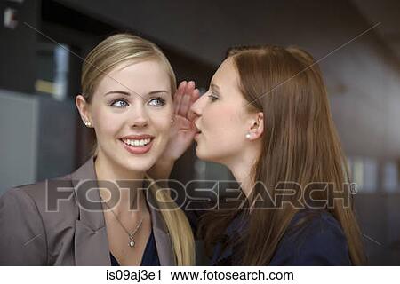 Stock Image - Two young businesswomen whispering in office. Fotosearch