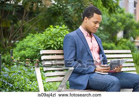 Young businessman sitting on park bench looking at digital tablet View Large Photo Image Stock Image - Young businessman sitting on park bench looking at digital tablet. Fotosearch