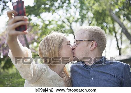 Stock Image - Young couple in park taking selfie whilst kissing. Fotosearch