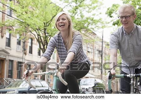 Picture - Young couple laughing whilst cycling along street. Fotosearch