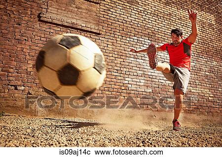 Stock Image - Young man kicking soccer ball on wasteland. Fotosearch