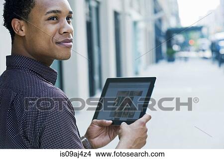 Picture - Young man on city street using digital tablet and looking over shoulder. Fotosearch