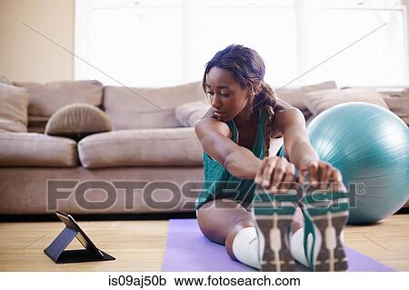 Stock Image - Young woman exercising on sitting room floor whilst looking at digital tablet. Fotosearch