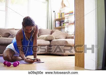 Stock Image - Young woman exercising on sitting room floor whilst looking at smartphone. Fotosearch