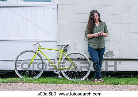 Stock Image - Young woman leaning against house texting on smartphone. Fotosearch
