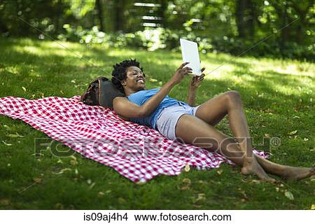 Picture - Young woman lying in park looking up at digital tablet. Fotosearch