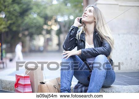 Young woman shopper sitting on city steps chatting on smartphone View Large Photo Image Picture - Young woman shopper sitting on city steps chatting on smartphone. Fotosearch