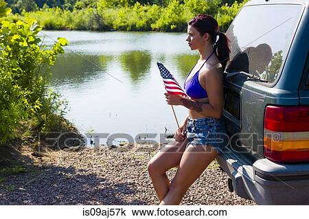 Stock Image - Young woman with american flag leaning against off road vehicle, Delaware Canal State Park, New Hope, Pennsylvania, USA. Fotosearch
