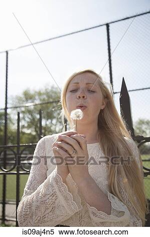 Stock Photography - Young woman with eyes closed blowing dandelion clock in park. Fotosearch