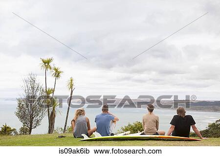 Stock Image - Four young adult surfer sitting on grass looking away to sea. Fotosearch