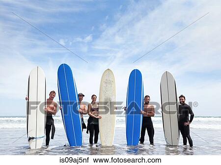 Stock Image - Group portrait of male and female surfer friends standing in sea with surfboards. Fotosearch