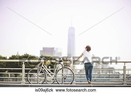 Stock Image - Mid adult female cyclist gazing at city from riverside, New York City, USA. Fotosearch