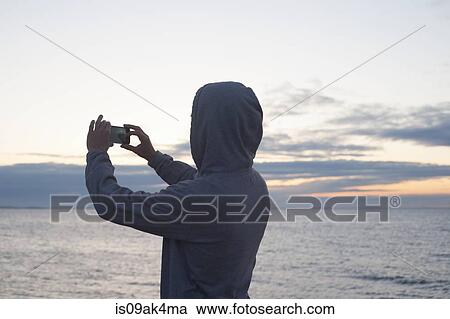 Stock Image - Mid adult man in hooded top photographing sea with smartphone, Gloucester, Massachusetts, USA. Fotosearch