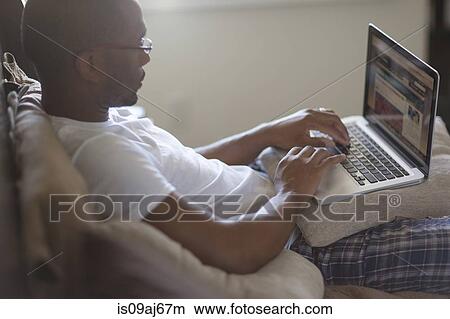Picture - Mid adult man, lying on bed, using laptop. Fotosearch