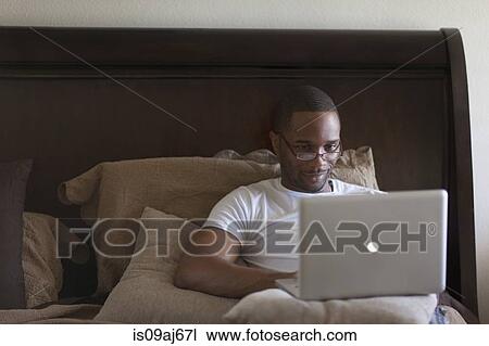 Stock Image - Mid adult man, lying on bed, using laptop. Fotosearch