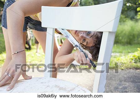 Stock Photo - Mid adult woman bending forward to paint and restore chair in garden. Fotosearch