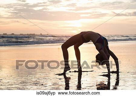 Stock Photography - Mid adult woman bending over backwards on beach at sunset, Nosara, Guanacaste, Costa Rica. Fotosearch