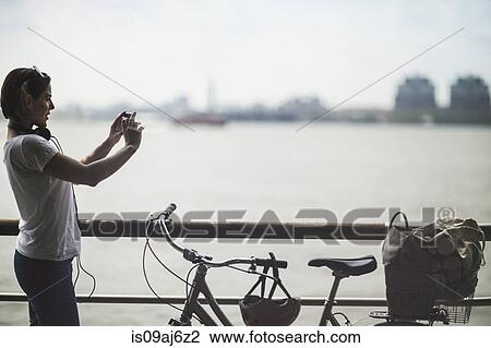 Stock Image - Mid adult woman cyclist photographing with smartphone at riverside, New York City, USA. Fotosearch