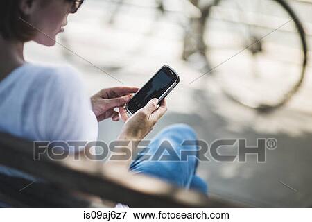 Mid adult woman cyclist sitting on bench looking at smartphone View Large Photo Image Stock Photo - Mid adult woman cyclist sitting on bench looking at smartphone. Fotosearch