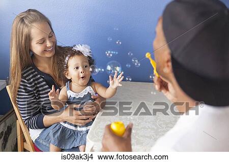 Picture - Mother and father playing with young daughter, father blowing bubbles. Fotosearch
