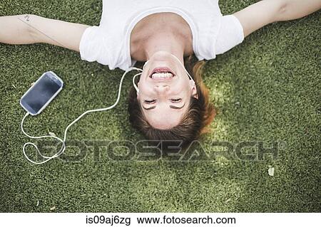 Stock Image - Overhead portrait of mid adult woman listening to earphones in park. Fotosearch