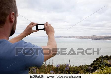 Stock Image - Rear cropped view of young man photographing sea with smartphone. Fotosearch