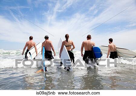 Stock Image - Rear view of a group of male and female surfer friends wading into sea with surf boards. Fotosearch