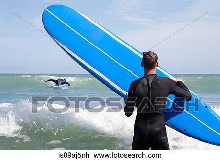 Stock Image - Rear view of male surfer with surfboard watching friend surfing. Fotosearch