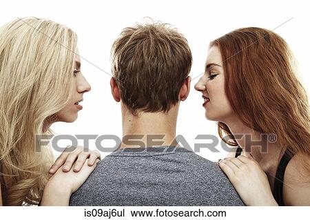 Stock Image - Studio portrait of two young woman whispering into young man's ears. Fotosearch