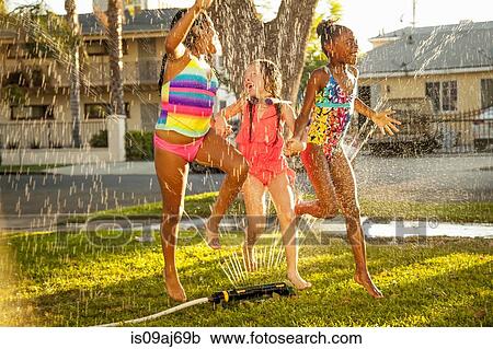 Stock Image - Three girls running and jumping in garden sprinkler. Fotosearch