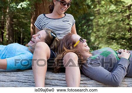Stock Image - Three women relaxing and laughing on tree trunk in forest. Fotosearch