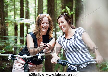 Stock Photo - Two women mountain bikers looking at smartphone in forest. Fotosearch