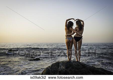 Stock Image - Two young women standing on rock in ocean, rear view. Fotosearch
