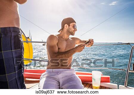 Stock Photo - Young man sitting on boat using smartphone. Fotosearch