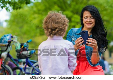 Young mother photographing son on smartphone in park View Large Photo Image Picture - Young mother photographing son on smartphone in park. Fotosearch