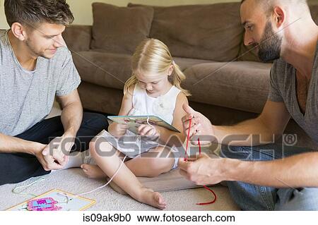 Stock Image - Male couple and daughter threading picture books on sitting room floor. Fotosearch