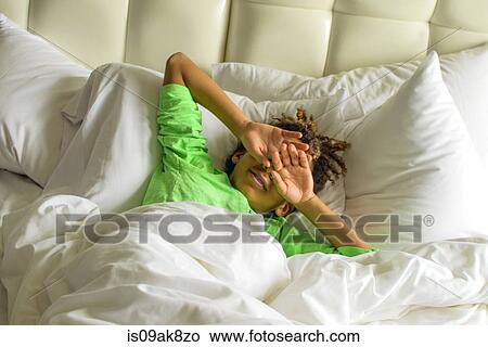 Picture - Sleepy boy lying in bed shielding face with hands. Fotosearch
