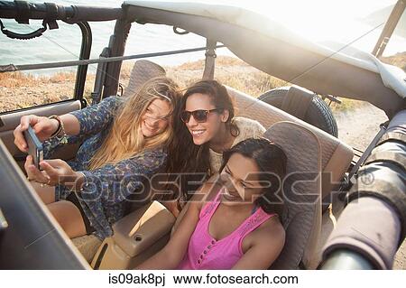 Stock Image - Three young women taking selfie on smartphone in jeep at coast, Malibu, California, USA. Fotosearch
